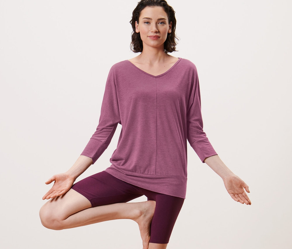 Frau in Baumstellung auf einer Yogamatte. Sie trägt ein Yogashirt in Mauve.