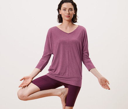 Frau in Baumstellung auf einer Yogamatte. Sie trägt ein Yogashirt in Mauve.