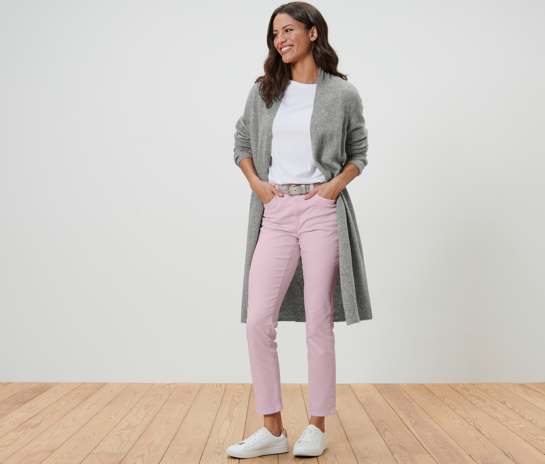 Eine Frau in einem offenen Longcardigan, einem weißen T-Shirt, einer rosafarbenen Hose und weißen Turnschuhen steht auf einem Holzboden.