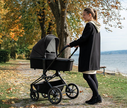 Eine Frau schiebt einen schwarzen Osann Kombi-Kinderwagen »Vamos« auf einem Weg mit Herbstlaub.