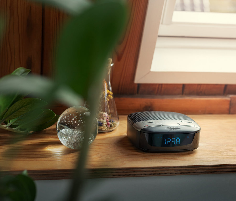 Auf einem Holzregal steht ein schwarzer Philips Radiowecker »TAR3505« mit einer Anzeige, die die Uhrzeit 12:38 anzeigt, neben einer Glaskugel und einer Vase mit Blumen.