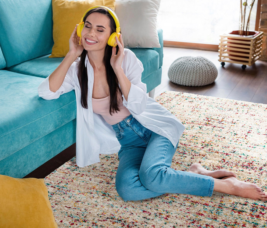 Frau sitzt auf einem beigen Teppich »Relic« mit gedämpften Pastelltönen und hört Musik mit gelben Kopfhörern.