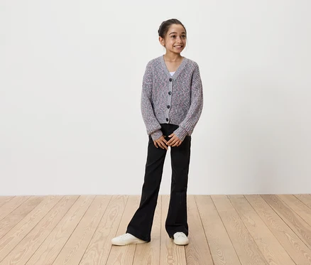 Lächelndes Mädchen in grauem Strickcardigan mit Knöpfen, schwarzer Schlaghose und weißen Turnschuhen steht auf einem Holzboden vor einer weißen Wand.
