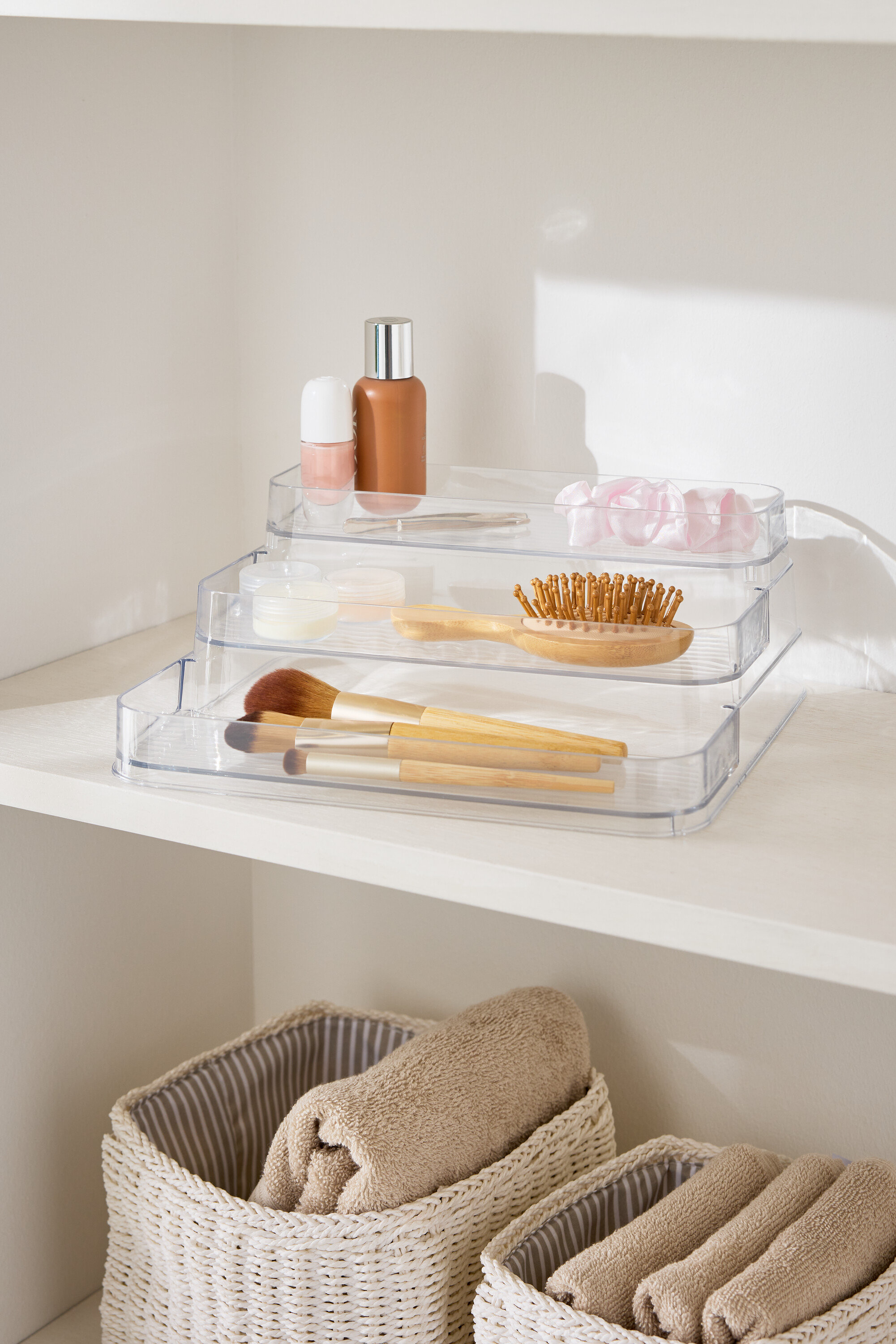 Dreistufiger, durchsichtiger Make-up-Organizer auf einem weißen Regal neben Körben mit Handtüchern.