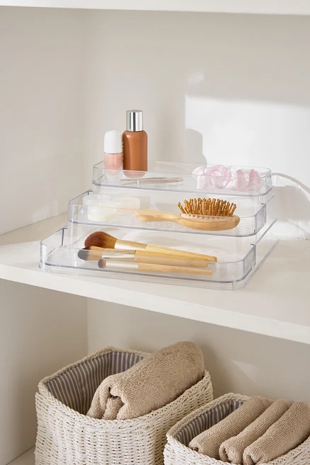 Dreistufiger, durchsichtiger Make-up-Organizer auf einem weißen Regal neben Körben mit Handtüchern.