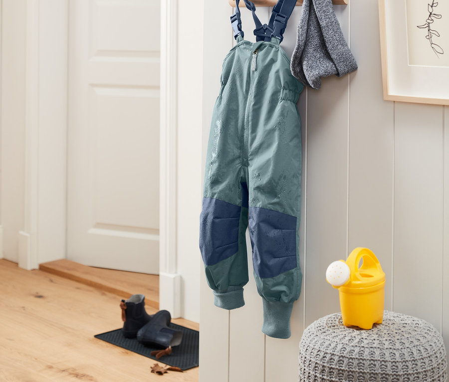 Regenhose aus recyceltem Material mit Mitwachsfunktion hängt an der Wand neben einem grauen Tuch.