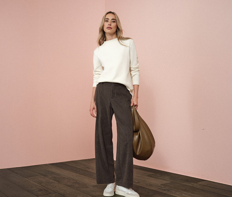 Frau steht und hält eine Tasche, sie trägt eine Street One Hose Straight Leg, einen weißen Pullover und Turnschuhe.