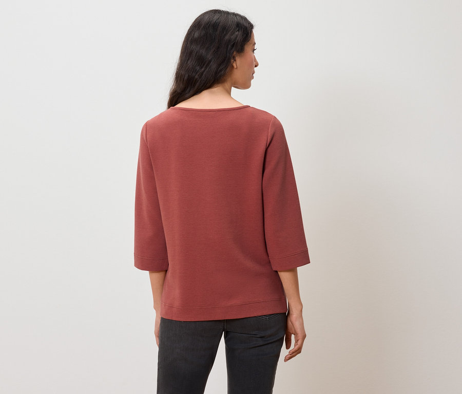 Rückansicht einer Frau mit dunklen Haaren, die ein rotbraunes Sweatshirt mit Dreiviertelärmeln und dunkle Jeans trägt.