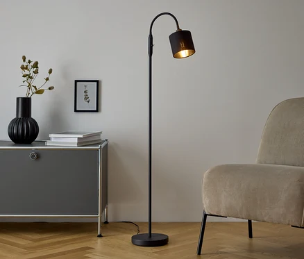 Eine schwarze Stehlampe steht neben einer grauen Kommode, auf der eine Vase und Bücher stehen. Neben der Kommode steht ein beiger Sessel.