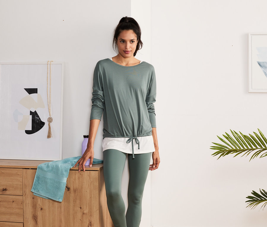 Eine Frau steht in einem Raum und trägt ein grün-weißes 2-in-1-Sportshirt.