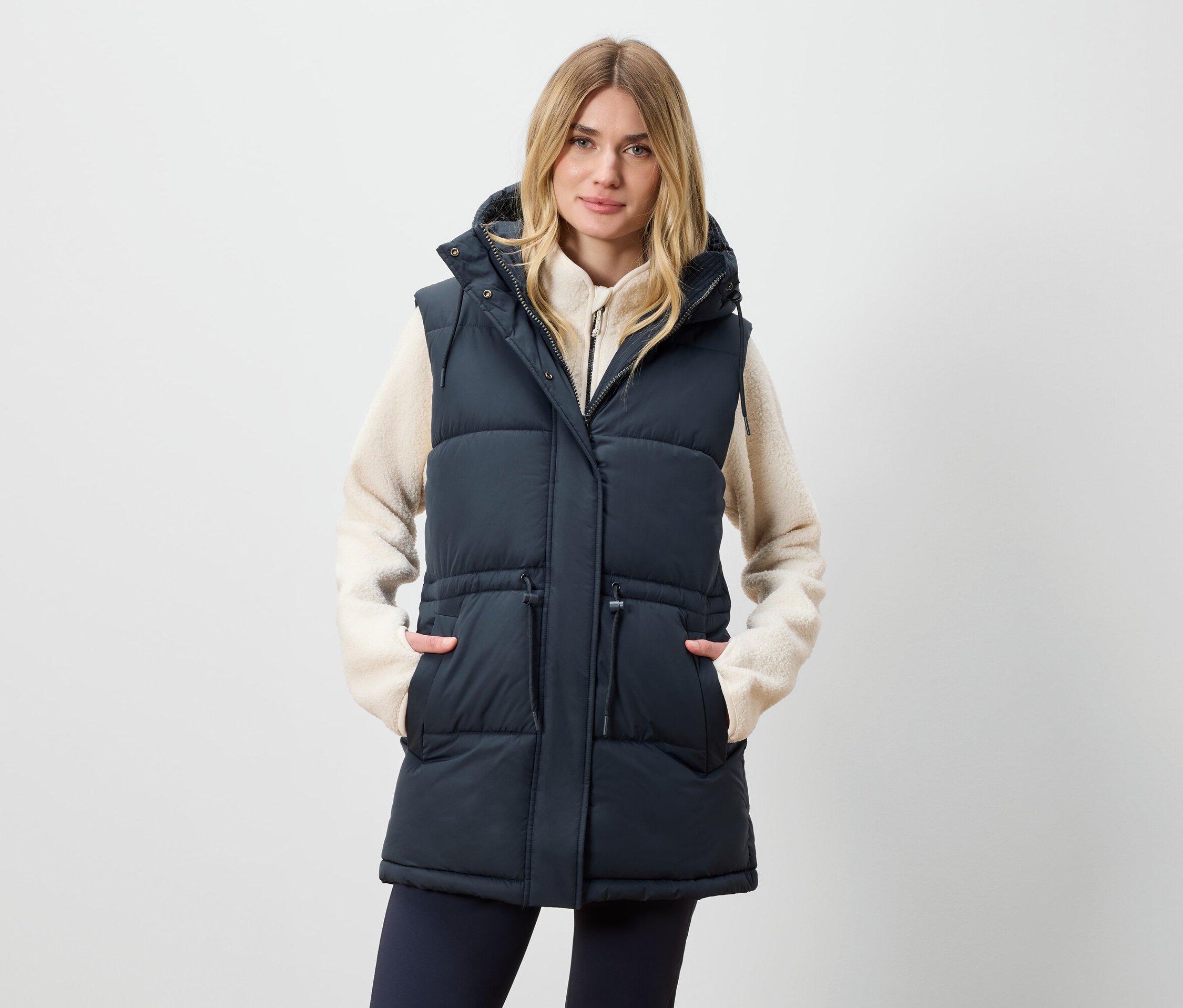 Frau trägt eine lange Steppweste mit Kapuze und hat die Hände in den Taschen.