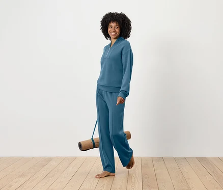Lächelnde Frau mit Afro-Frisur geht barfuß und trägt eine Yogamatte, während sie ein blaues Trainingsanzug-Set trägt.