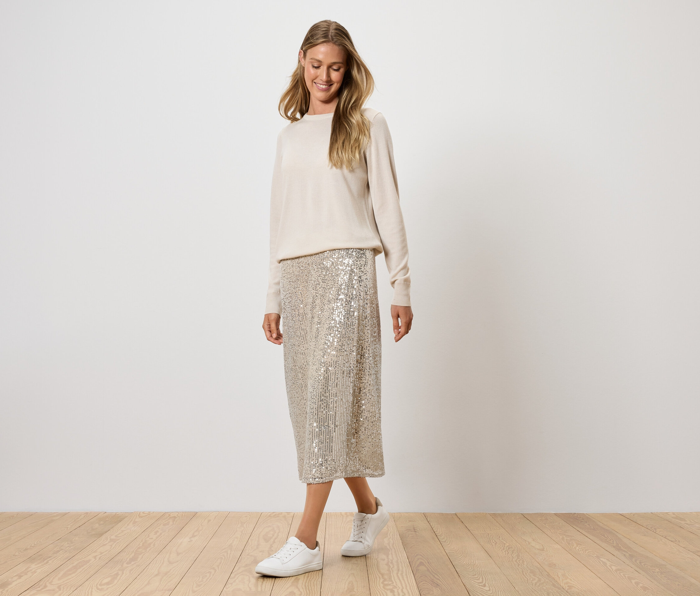 Frau posiert in cremefarbenem Pullover, Paillettenrock und weißen Turnschuhen.