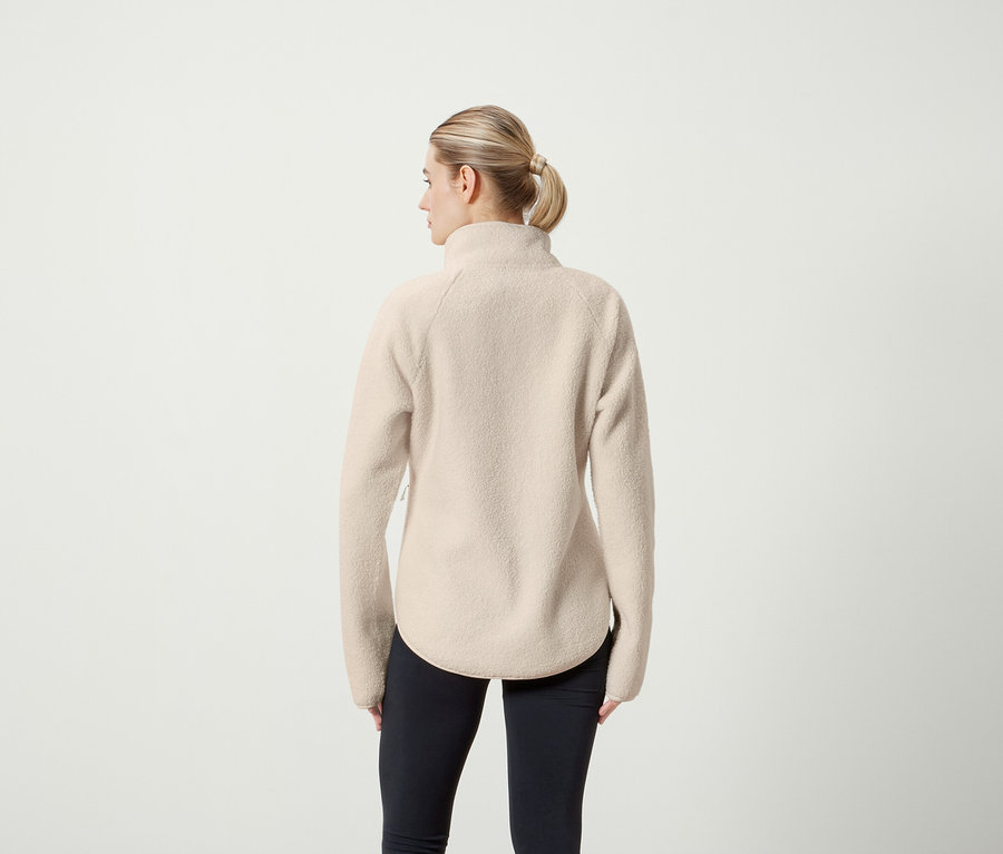 Von hinten ist eine Frau mit blonden, zu einem Pferdeschwanz zusammengebundenen Haaren zu sehen, die eine beige Fleecejacke und schwarze Leggings trägt.