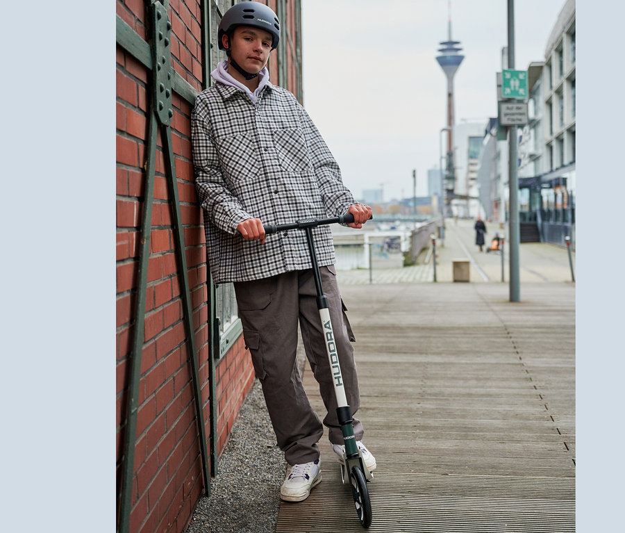 Ein Jugendlicher mit kariertem Hemd und grauem Helm steht im Freien an einer Backsteinmauer neben einem grau-weißen HUDORA BigWheel® 205 Scooter.