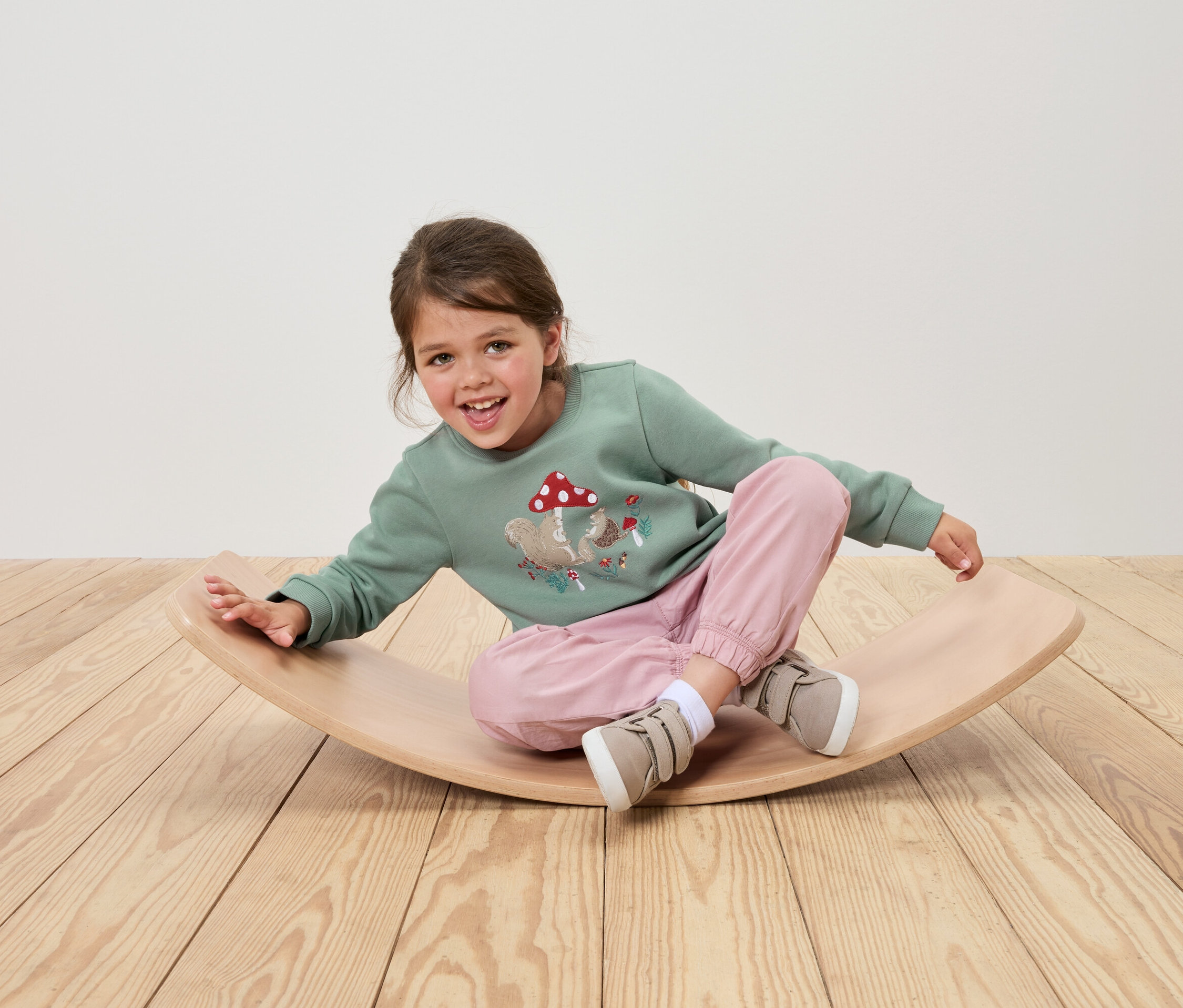 Ein Mädchen sitzt auf einem hölzernen Balance Board und trägt ein grünes Sweatshirt mit Eichhörnchen- und Pilzaufdruck, eine rosa Hose und beige Schuhe.