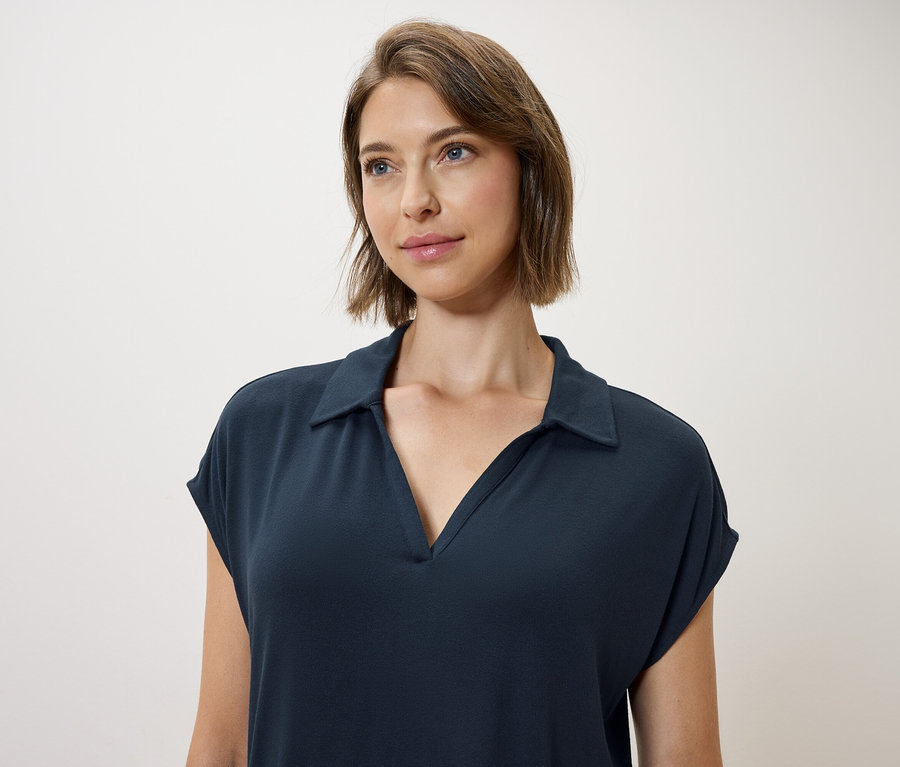 Frau mit braunen Haaren und blauen Augen trägt ein dunkelblaues Polokleid.