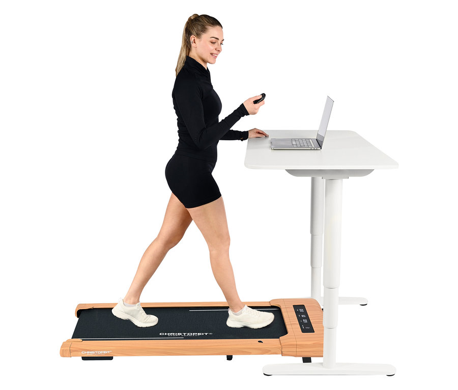 Frau geht auf Walking-Pad »4.2 Holzoptik« unter einem weißen Schreibtisch mit Laptop.