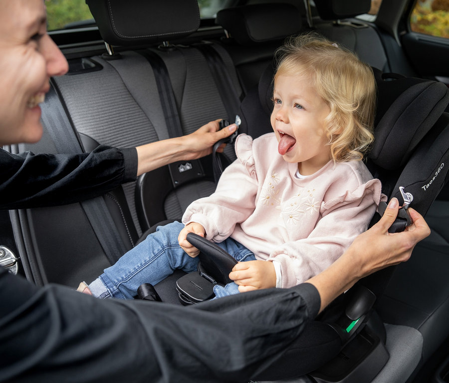 Kleinkind sitzt in schwarzem Osann Kinder-Autositz »Taos 360« und streckt Zunge heraus, während eine erwachsene Person es anschnallt.