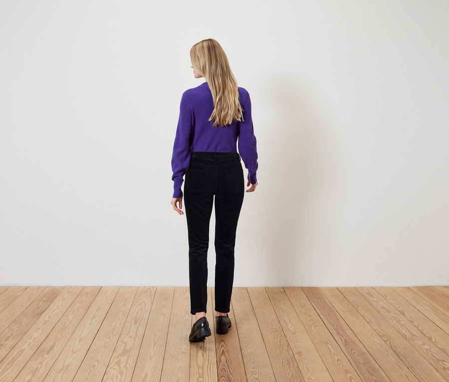 Eine Frau mit blonden Haaren steht mit dem Rücken zur Kamera und trägt einen violetten Pullover und eine schwarze Hose.