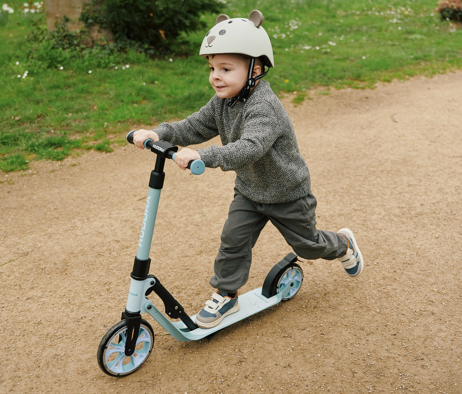 Junge mit grauem Pullover und Hose fährt mit blauem HUDORA BigWheel 180 Advanced Junior Scooter auf einem Feldweg.