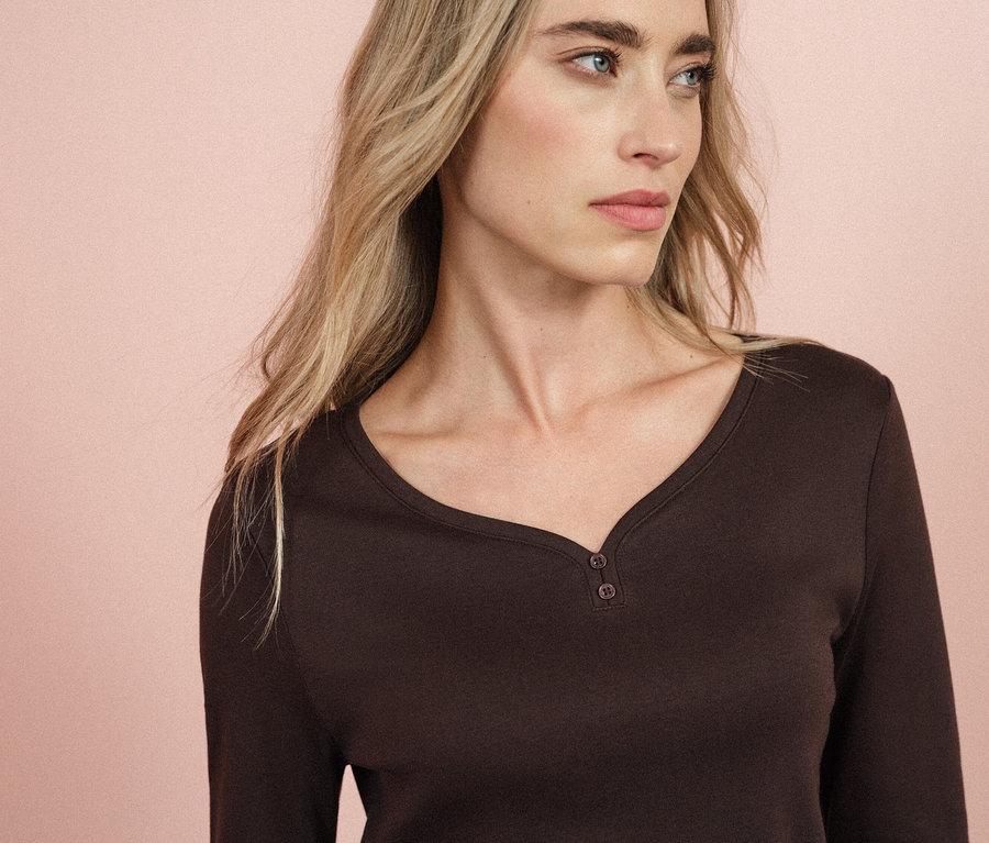 Blonde Frau mit blauen Augen trägt ein braunes Langarmshirt.