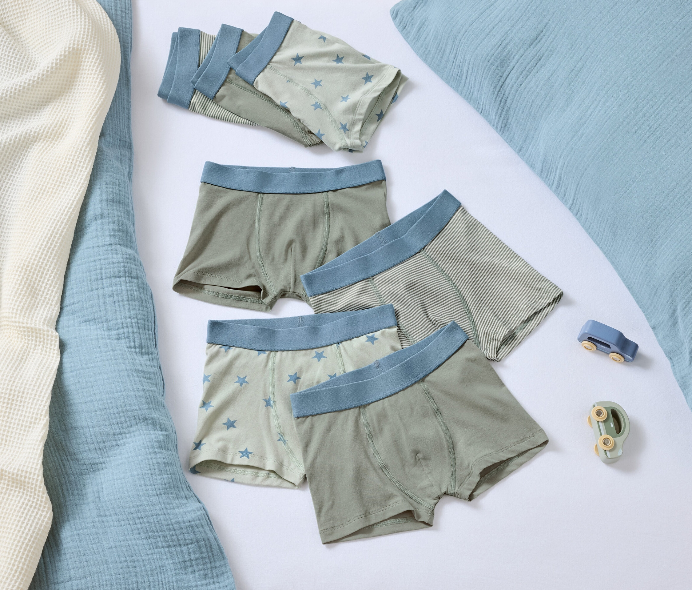 Fünf Boxershorts mit blauem Bund liegen auf einem weißen Hintergrund neben einem blauen Kissen und zwei kleinen Spielzeugen.