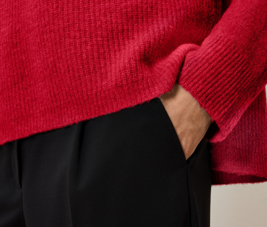 Detailaufnahme eines roten Pullovers und einer schwarzen Hose mit einer Hand in der Tasche.