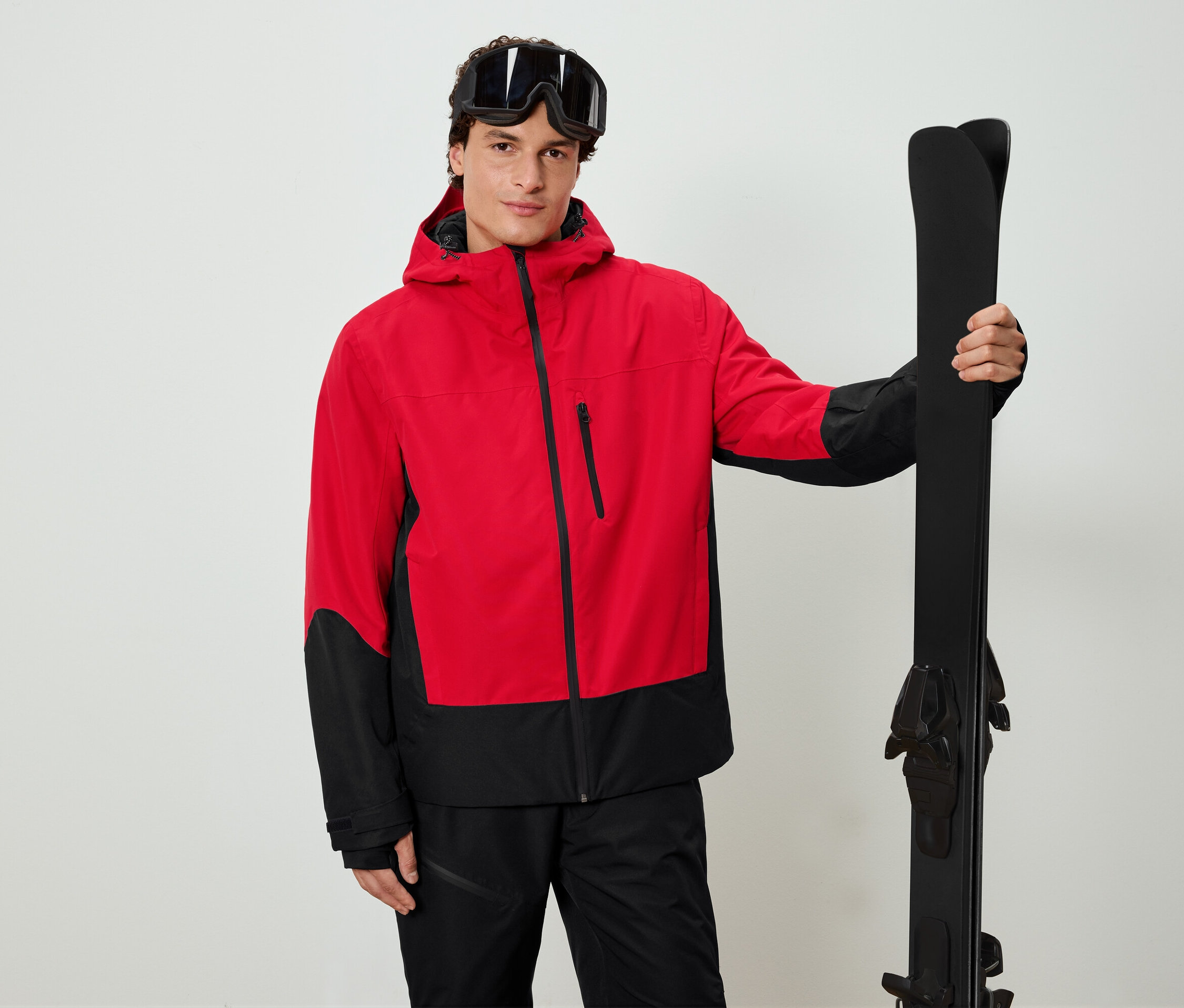 Mann mit rot-schwarzer Jacke hält schwarze Ski und trägt eine Skibrille.