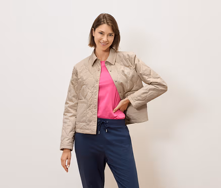 Eine Frau mit kurzem braunem Haar posiert vor einer cremefarbenen Wand in einer beige gesteppten Jacke, einem pinkfarbenen T-Shirt und blauen Hosen.