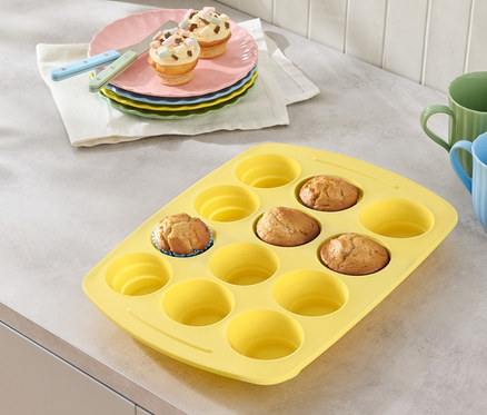 Gelbe Muffinform mit ein paar Muffins, Teller mit Muffins und Gläser auf einer Arbeitsfläche.