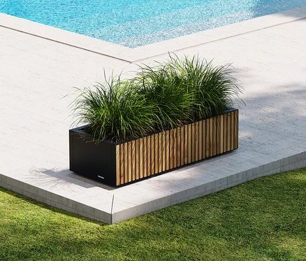 Ein länglicher Pflanzkasten mit grünen Gräsern steht auf einer Terrasse neben einem Pool.