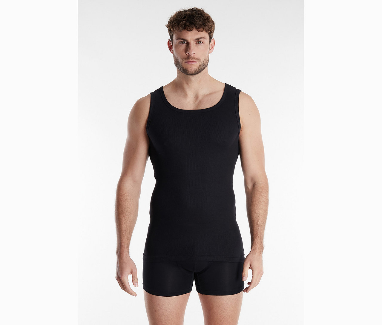 Mann mit lockigem Haar trägt ein schwarzes Tanktop und Boxershorts vor einem weißen Hintergrund.