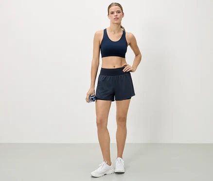 Frau steht mit der Hand auf der Hüfte, trägt einen marineblauen Sport-BH, Shorts und weiße Turnschuhe und hält eine Wasserflasche.