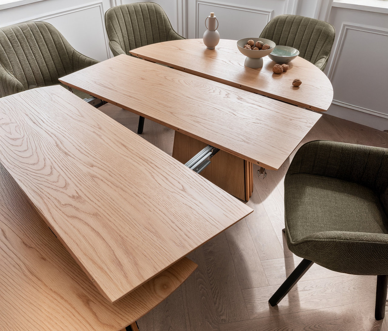 Ausgezogener Holztisch mit vier grünen Stühlen, einer Vase und einer Schale mit Nüssen.