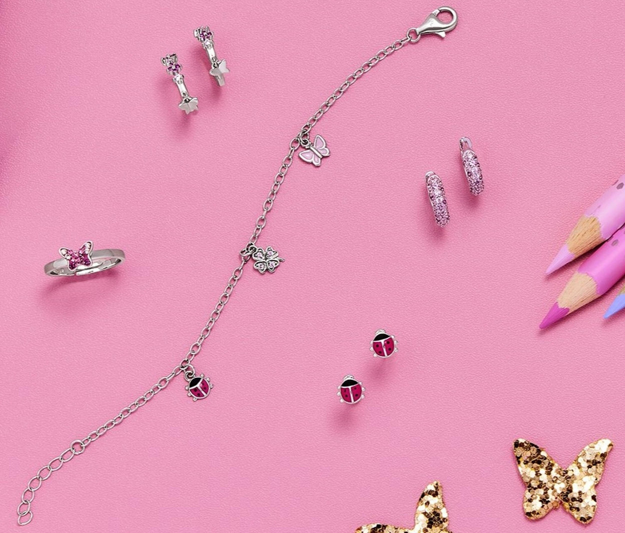Auf rosa Untergrund liegen Ohrschmuck »Zirkonia« aus 925er Silber, Armband mit Anhängern, Ring und Ohrstecker. Daneben liegen Stifte und Schmetterlinge.