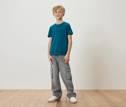Ein blonder Junge steht in einem türkisfarbenen T-Shirt, grauen Cargohosen und weißen Turnschuhen vor einer weißen Wand.