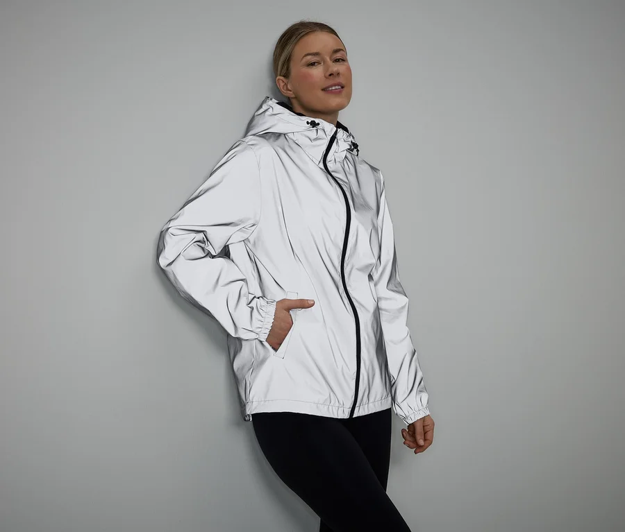 Frau mit Hand in Tasche, trägt reflektierende Jacke mit Kapuze und schwarzen Leggings vor grauem Hintergrund.