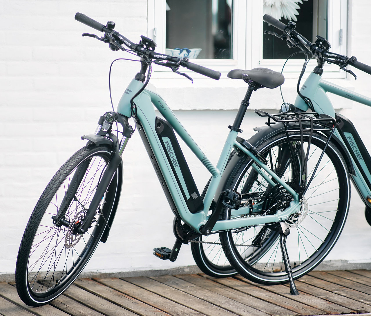 Zwei FISCHER Trekking E-Bikes VIATOR 2.1 ACTIVE mit Diamant-Rahmen stehen vor einer weißen Hauswand auf einem Holzsteg.