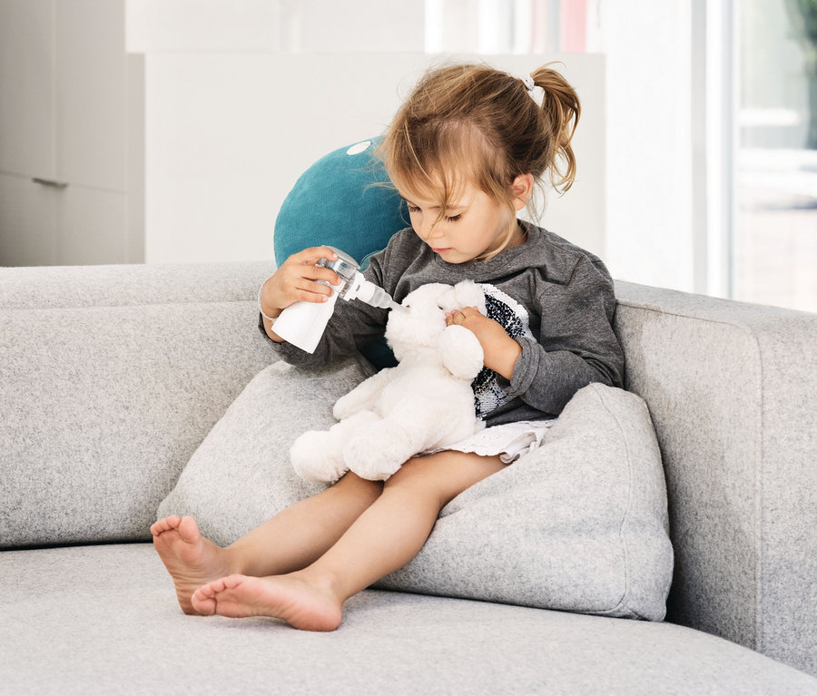 Mädchen inhaliert mit dem Beurer Inhalationsgerät »IH57« einen Teddybären auf einem grauen Sofa.