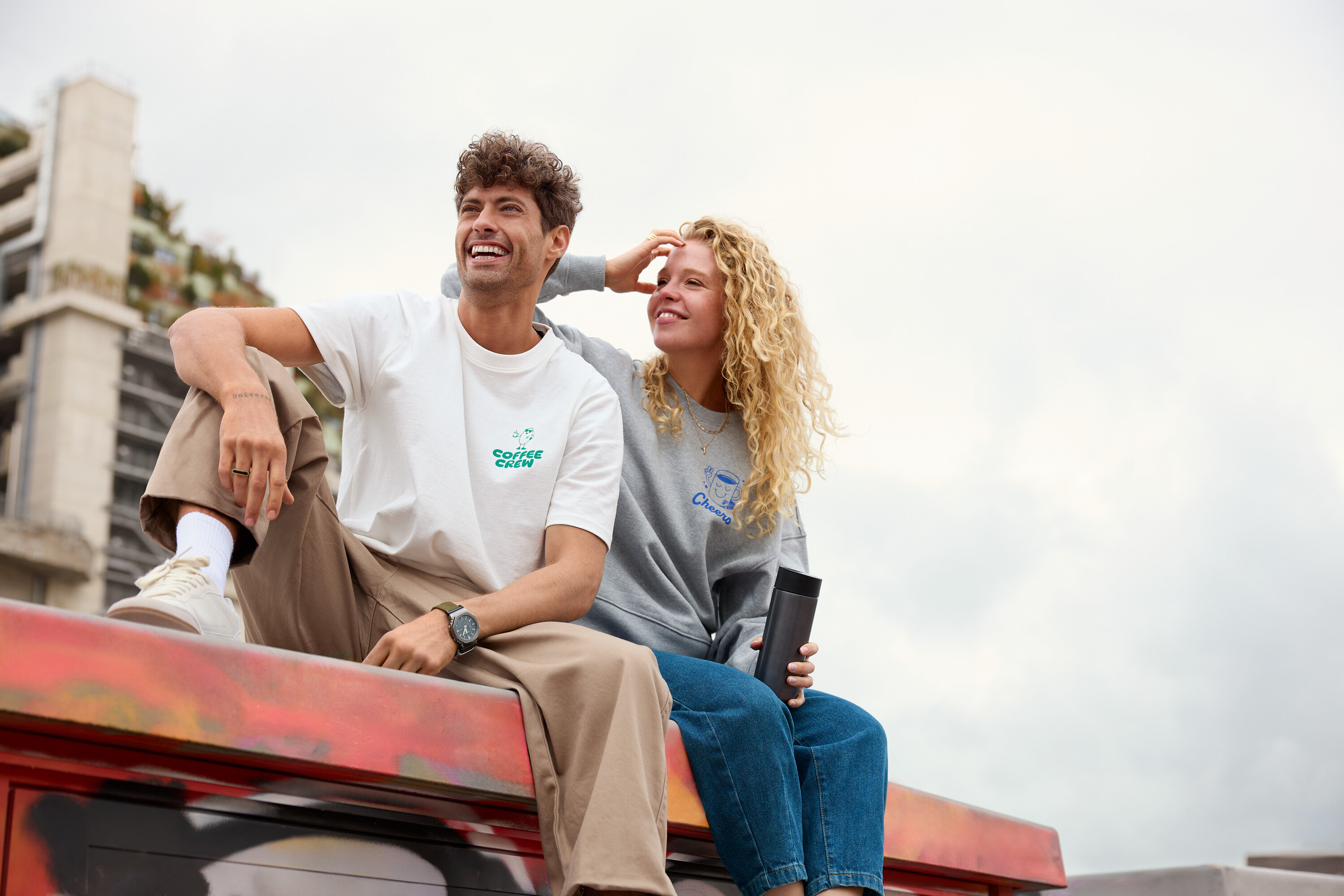 Ein Mann mit lockigem Haar und eine Frau mit lockigem Haar sitzen auf einer Mauer. Der Mann trägt ein weißes T-Shirt mit der Aufschrift "Coffee Crew" und die Frau einen grauen Pullover mit der Aufschrift "Cheers".