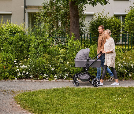 Ein Paar schiebt einen grauen Kinderwagen auf einem Gehweg neben einem bewachsenen Garten.
