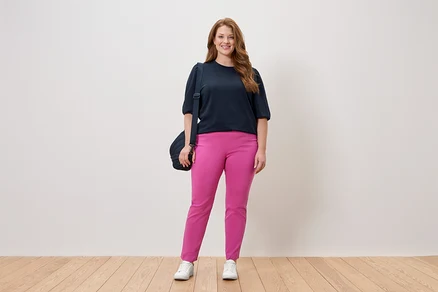 Frau steht in marineblauem Oberteil und pinkfarbener Hose mit einer Umhängetasche vor einer weißen Wand.