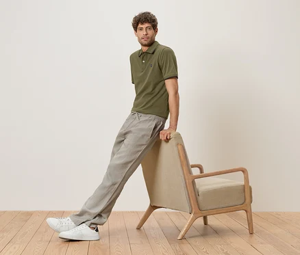 Ein Mann lehnt an einem beigen Sessel, trägt ein olivgrünes Poloshirt, graue Leinenhose und weiße Sneaker.