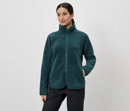 Frau mit Zopf trägt eine grüne Fleecejacke mit Reißverschluss und schwarze Hose, eine Hand in der Jackentasche.