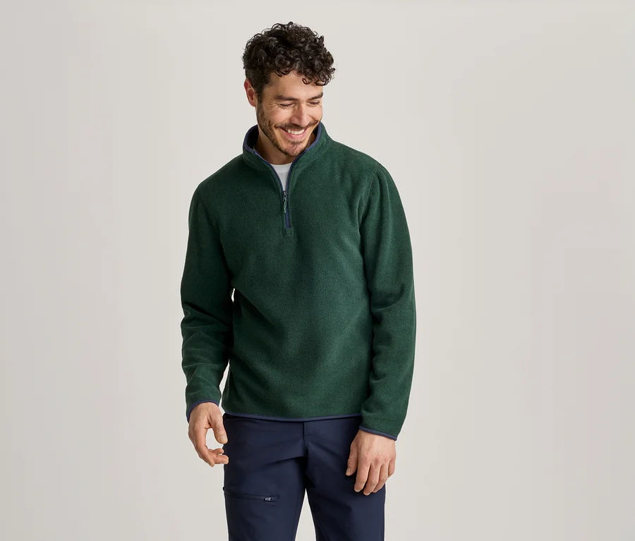 Ein lächelnder Mann mit dunklem Haar und Bart trägt ein dunkelgrünes Fleece-Oberteil mit Viertelreißverschluss und dunkelblauer Hose.