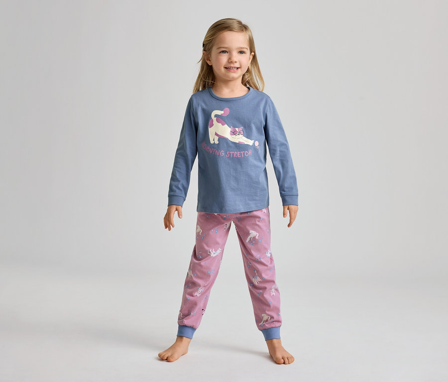 Ein junges Mädchen steht barfuß und lächelt in einem blauen Langarmshirt mit einer streckenden Katze und rosa Hosen mit Einhornmuster.