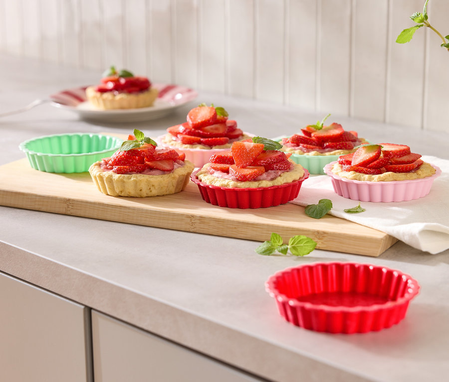 Mehrere Erdbeertörtchen in bunten Förmchen auf einem Holzbrett mit Minze garniert.