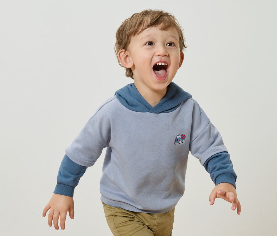 Ein kleiner Junge mit blonden Locken und offenem Mund trägt einen hellblauen Kapuzenpullover mit einer kleinen Auto-Applikation und hellbraune Hosen vor einem weißen Hintergrund.