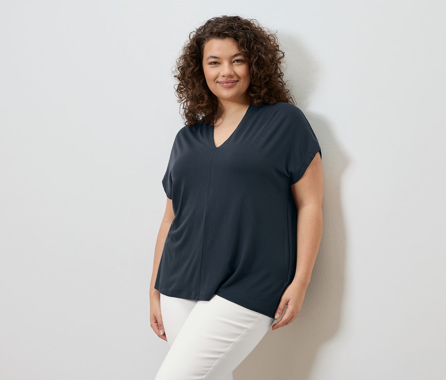 Eine Frau mit lockigem Haar steht vor einer weißen Wand und trägt ein marineblaues T-Shirt und eine weiße Hose.